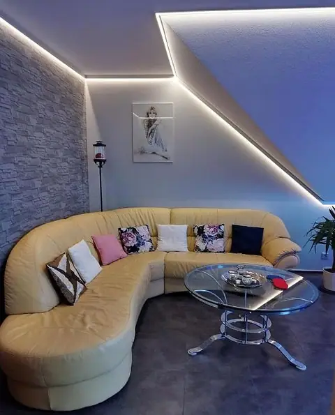 Wohnzimmer mit hellem Leder-Ecksofa, Glastisch und LED-Lichtband unter der Dachschräge