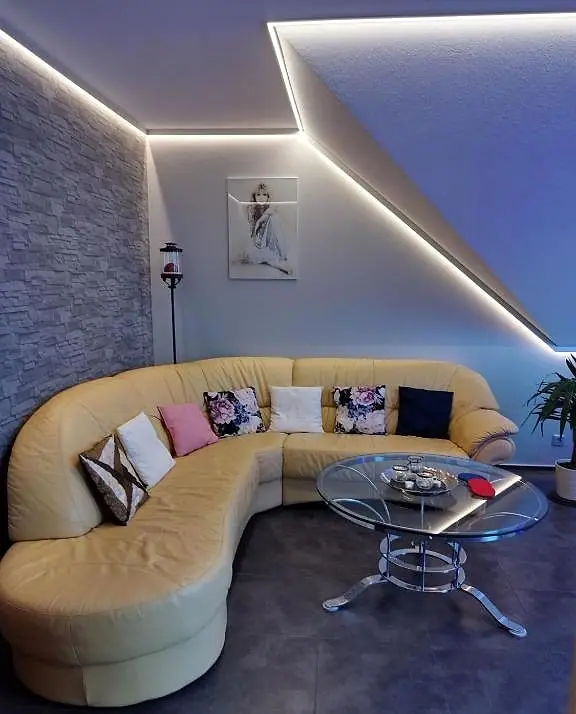 Wohnzimmer mit hellem Leder-Ecksofa, Glastisch und LED-Lichtband unter der Dachschräge